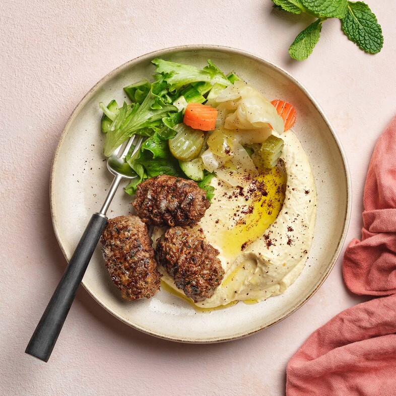 Kryddiga köftebiffar serverade med hummus, sallad och turkiska pickles på ett fat.
